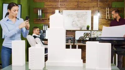 Young female architect taking photos of building models. Bearded architects working in the background.