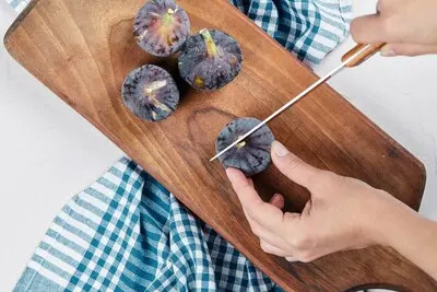 Hand cutting a fig on wooden cutting board with a blue tablecloth.